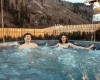 Dwie osoby relaksują się w zewnętrznym jacuzzi z drewnianym płotem i górami w tle.
