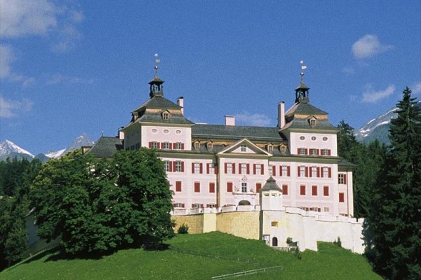 Historyczny zamek na wzgórzu otoczony drzewami z górami w tle.
