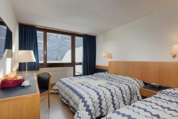 Pokój hotelowy z dwoma łóżkami, biurkiem i widokiem z okna na zaśnieżone góry.