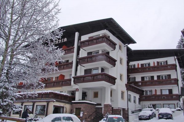 Czteropiętrowy hotel z ośnieżonymi balkonami i samochodami zaparkowanymi przed nim.