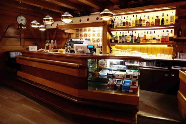 Drewniany bar z podświetlanymi półkami pełnymi butelek alkoholu i witryną ze słodyczami.