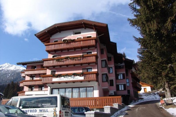 Górski hotel z drewnianymi balkonami i białym samochodem dostawczym na pierwszym planie, ośnieżone góry w tle.