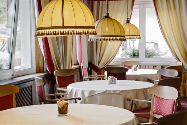Przytulna restauracja z okrągłymi stołami, czerwonymi krzesłami i zasłonami, oświetlona dużymi lampami.