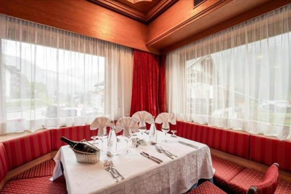 Stół dla sześciu osób z białym obrusem, elegancko nakryty w narożnej restauracji z czerwonymi zasłonami i dużymi oknami.