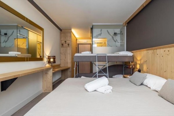 Pokój hotelowy z łóżkiem małżeńskim i piętrowym, drewnianymi meblami i dużym lustrem.