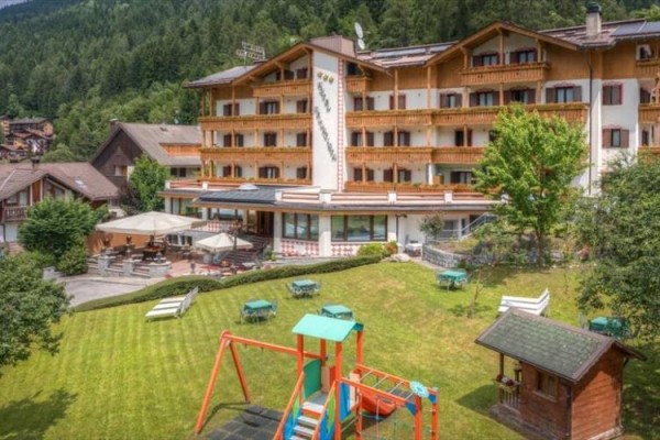 Hotel z tarasem i placem zabaw na trawniku otoczony lasem.