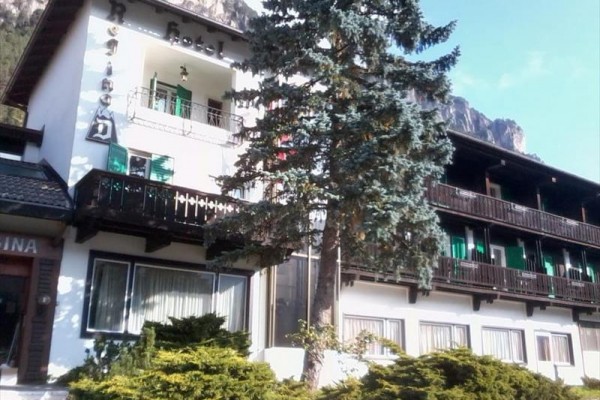 Hotel z balkonami i zielonymi okiennicami otoczony górami i drzewami.