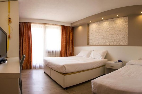 Nowoczesny pokój hotelowy z dwoma łóżkami, telewizorem i dużym oknem z pomarańczowymi zasłonami.