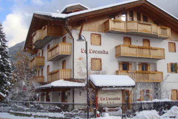 Dwupiętrowa górska chata La Locanda z drewnianymi balkonami pokryta śniegiem.