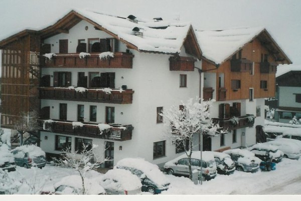 Ośnieżony alpejski hotel z balkonami i zaparkowanymi samochodami.