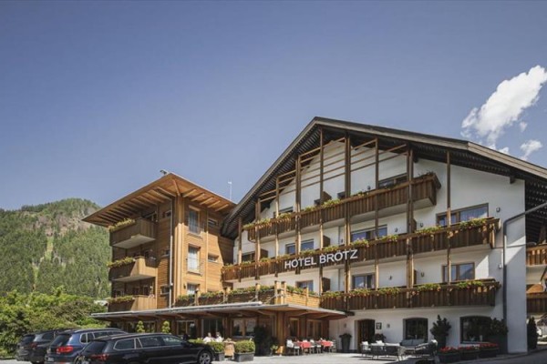 Budynek hotelu z drewnianymi balkonami i zaparkowanymi samochodami przed nim, w tle las i niebieskie niebo z chmurami.