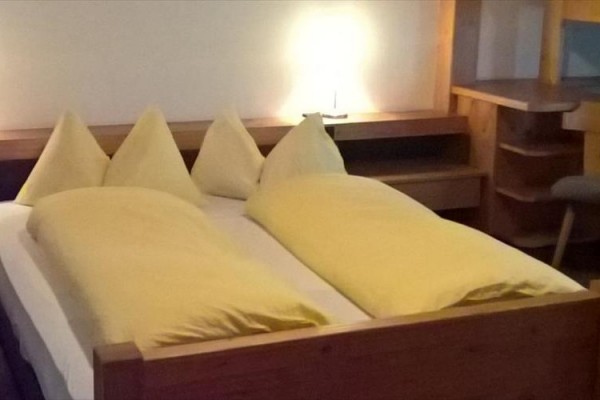Podwójne łóżko z drewnianą ramą i żółtą pościelą, obok stolika nocnego z włączoną lampką.