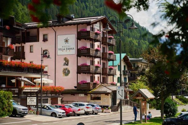 Różowy hotel Villa Emma z kwiatami na balkonach obok zaparkowanych samochodów w górskim regionie.