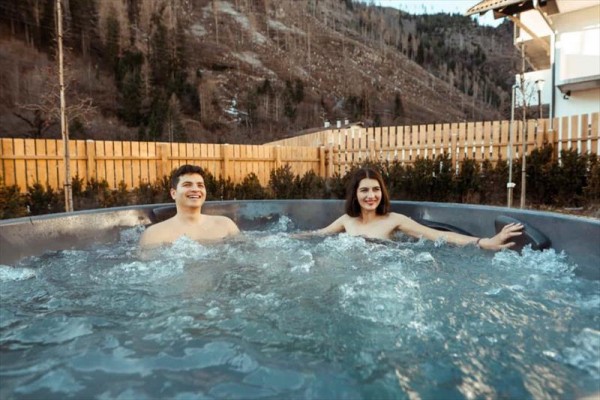 Dwie osoby relaksują się w zewnętrznym jacuzzi z drewnianym płotem i górami w tle.