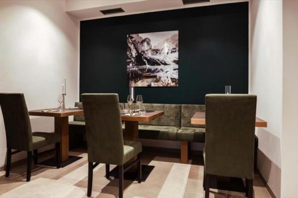 Restauracja z drewnianymi stołami i zielonymi krzesłami na tle ciemnej ściany z obrazem gór.