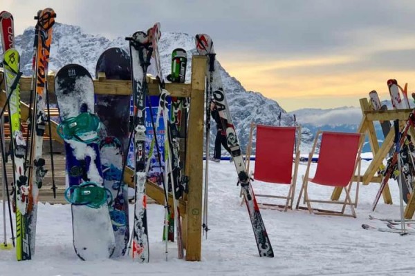 Narty i snowboardy oparte o stojaki na zaśnieżonym stoku z górami w tle.