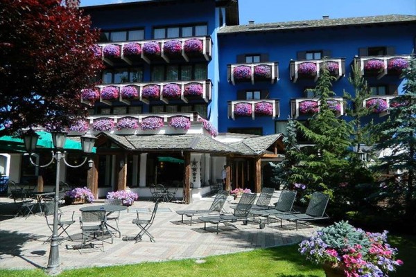 Niebieski budynek hotelu otoczony kwiatami na balkonach z tarasem wyposażonym w leżaki i stół.
