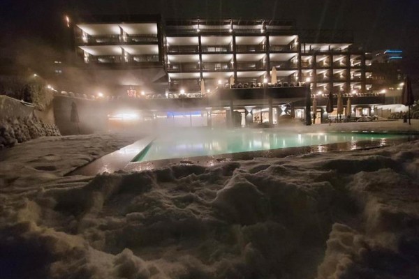 Oświetlony hotel z podgrzewanym basenem i śniegiem na pierwszym planie podczas ciemnej nocy.