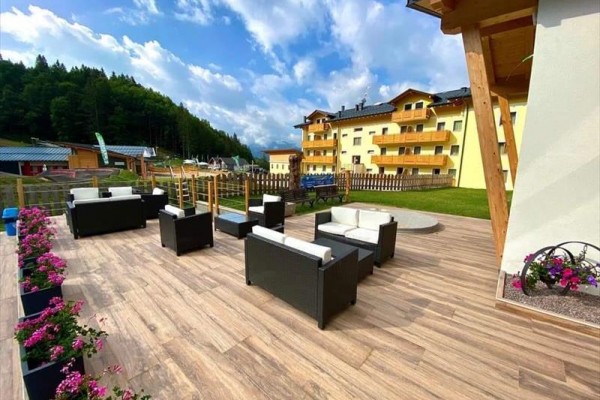 Taras z meblami ratanowymi, kwiatami i w tle żółty hotel przy lesie.