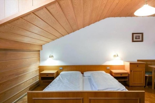 Sypialnia z drewnianą boazerią, podwójnym łóżkiem i stolikami nocnymi pod skośnym sufitem.  