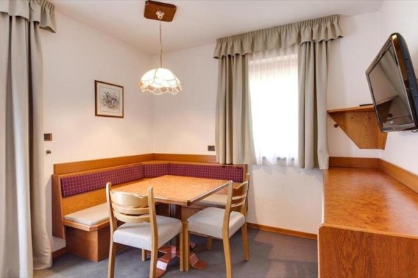 Kącik jadalniany z narożną ławką, drewnianym stołem i trzema krzesłami w pokoju z telewizorem na ścianie.