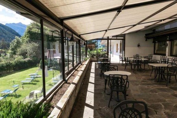 Zadaszony taras z metalowymi stołami i krzesłami obok ogrodu z leżakami.