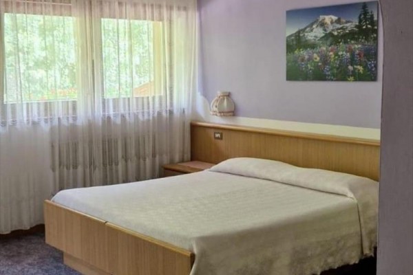 Prosto urządzona sypialnia z łóżkiem małżeńskim, obrazem góry na ścianie i zasłonami w oknach.