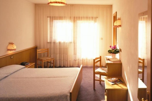 Jasny pokój hotelowy z łóżkiem, stolikami nocnymi, krzesłami i toaletką obok okna z firankami.