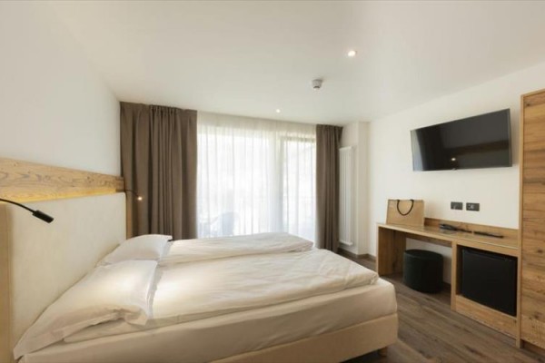 Nowoczesny pokój hotelowy z łóżkiem małżeńskim, telewizorem na ścianie i francuskimi oknami z ciemnymi zasłonami.