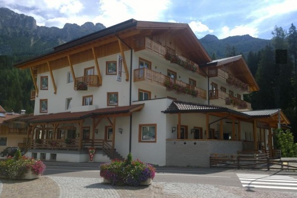 Trzypiętrowy hotel górski otoczony górami i lasem z kwiatami na balkonach.