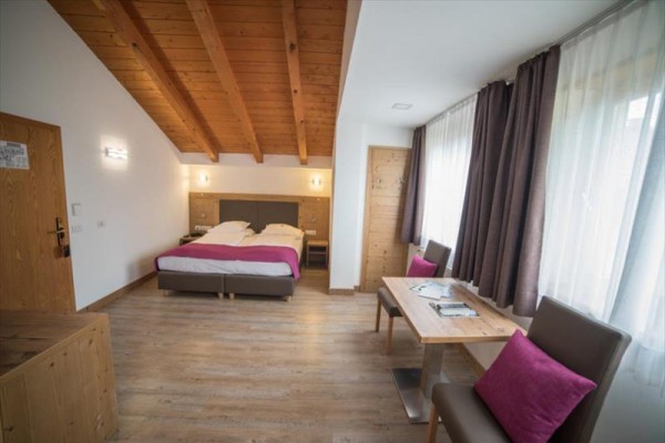 Pokój hotelowy z podwójnym łóżkiem, drewnianymi meblami i dużymi oknami z brązowymi zasłonami.