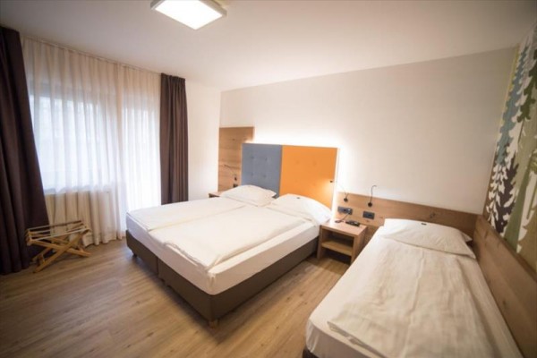 Pokój hotelowy z łóżkiem małżeńskim i pojedynczym, obok nich stolik nocny i zasłony przy oknie.