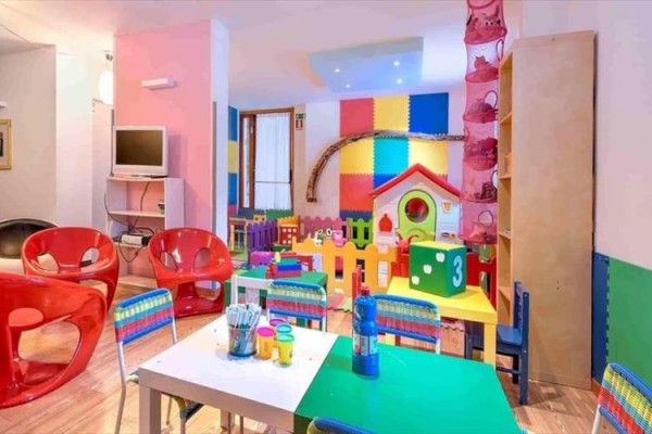 Sala zabaw dla dzieci z kolorowymi krzesłami, stołami, zabawkami i domkiem do zabaw.
