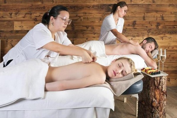 Dwie osoby leżą na stołach do masażu i są masowane w spa z drewnianą ścianą w tle.