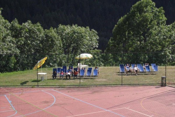 Leżaki z parasolem na trawniku obok pustego boiska sportowego otoczonego drzewami.