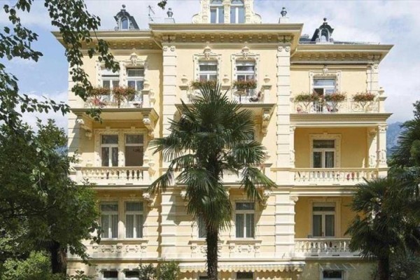 Żółty secesyjny budynek z balkonami, otoczony palmami i drzewami.