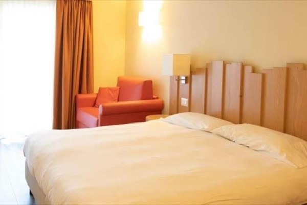 Jasny pokój hotelowy z podwójnym łóżkiem, czerwonym fotelem i drewnianym wezgłowiem łóżka.