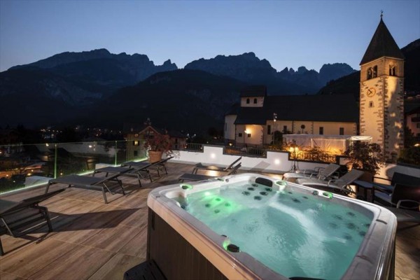 Zewnętrzne jacuzzi na tarasie z widokiem na góry i kościół o zmierzchu.