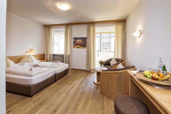 Jasny pokój hotelowy z podwójnym łóżkiem, stołem z owocami i fotelem przy oknie.