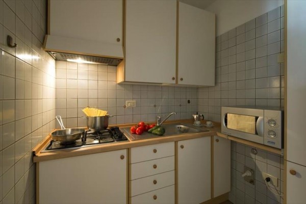 Mała kuchnia z kuchenką mikrofalową, kuchenką z garnkiem na makaron i zlewem z talerzem pomidorów i ogórka.