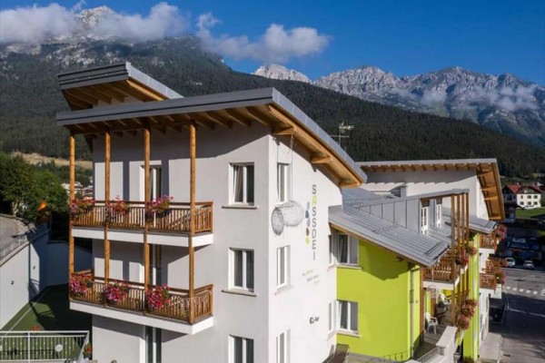 Nowoczesny hotel górski z zieloną i białą elewacją, w tle góry i pochmurne niebo.