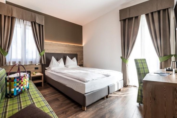 Nowocześnie urządzony pokój hotelowy z podwójnym łóżkiem, zielono-brązowymi zasłonami i telewizorem na ścianie.