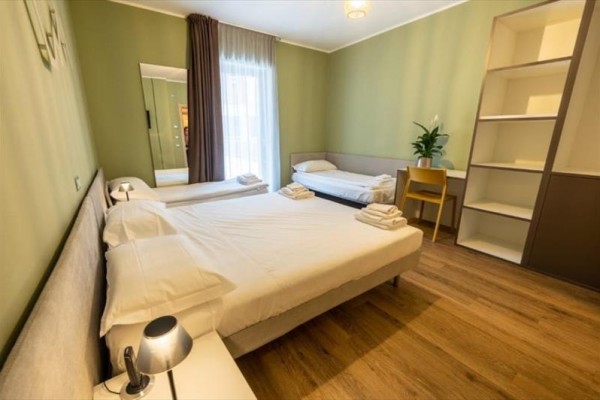 Pokój z dwoma łóżkami, biurkiem, otwartą półką i zasłonami przy oknie.