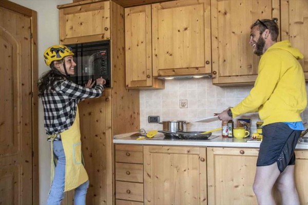 Dwie osoby gotują w drewnianej kuchni, jedna z nich ma kask podczas korzystania z mikrofalówki.