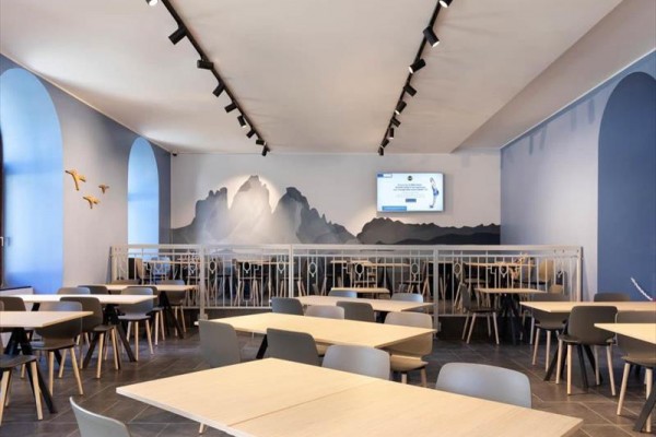 Nowoczesna restauracja z drewnianymi stołami, szarymi krzesłami i ściennym motywem gór.