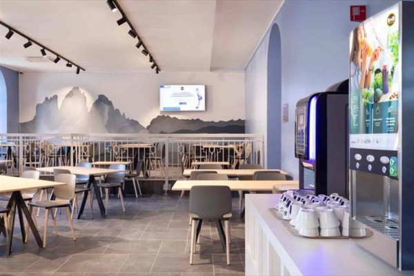 Nowoczesne wnętrze kawiarni z automatem do kawy i malowidłem gór na ścianie w tle.