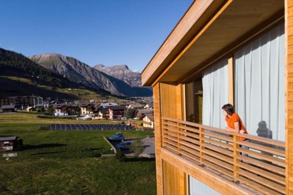 Osoba stoi na balkonie drewnianego domu z widokiem na pagórkowaty krajobraz.