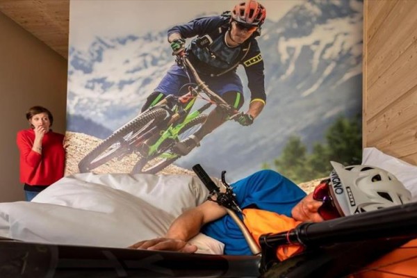 Mężczyzna śpi na łóżku obok plakatu rowerzysty, podczas gdy kobieta go zaniepokojona obserwuje.