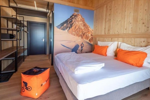 Nowoczesny pokój hotelowy z dużym łóżkiem, pomarańczowymi poduszkami i ścienną fotografią górskiej scenerii.