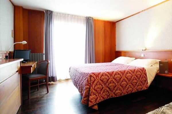 Pokój hotelowy z łóżkiem małżeńskim, biurkiem i światłem przenikającym przez zasłony.